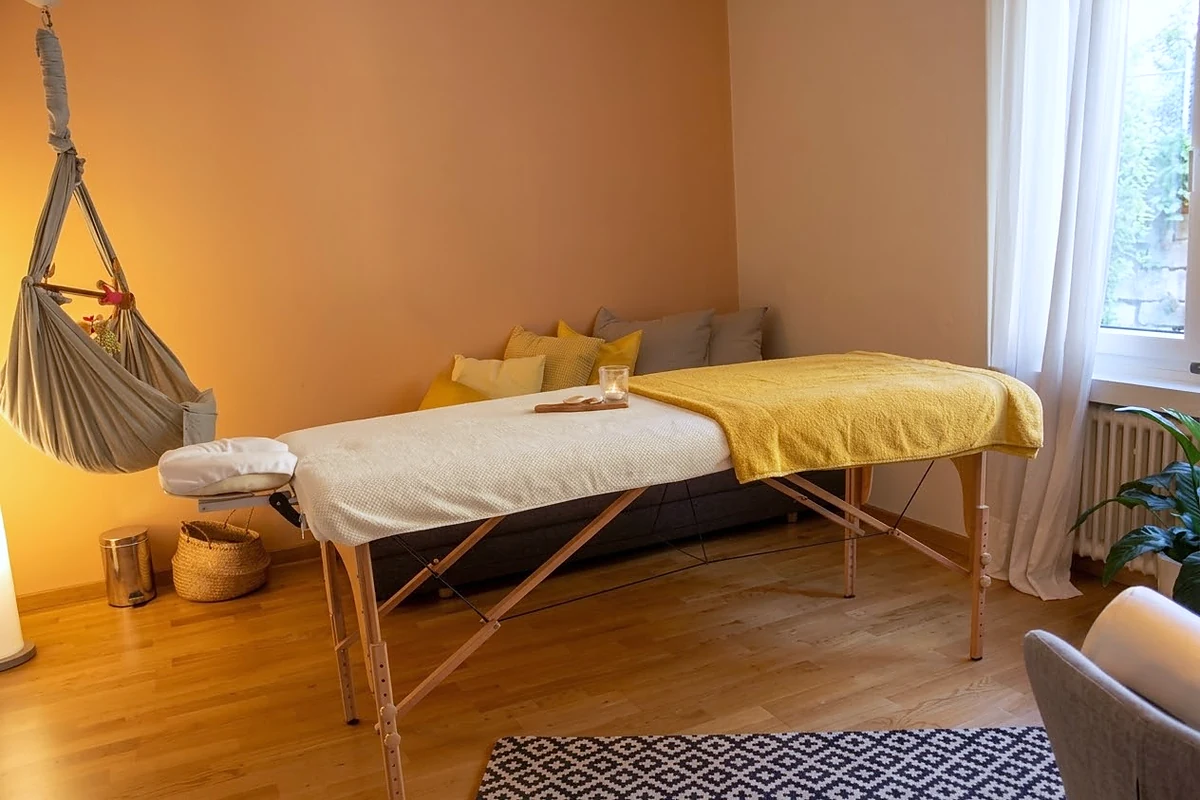 Der Behandlungsraum: hergerichtete Massageliege mit hellen Tüchern und gelber Decke, Kissen an der Wand, Tageslicht vom Fenster